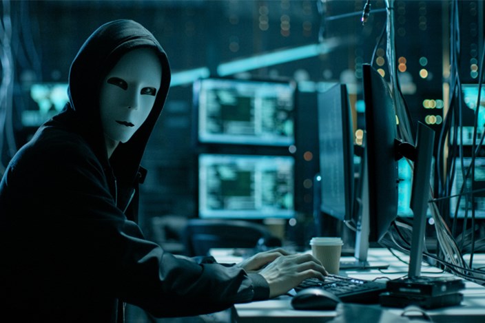 Warum Cyber-Sicherheit für Unternehmen Priorität haben sollte Hacker mit weißer Maske sitzt in einem abgedunkelten Raum mit vielen Monitoren.