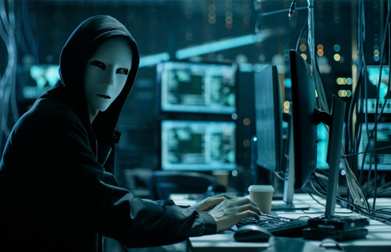 Warum Cyber-Sicherheit für Unternehmen Priorität haben sollte Hacker mit weißer Maske sitzt in einem abgedunkelten Raum mit vielen Monitoren.