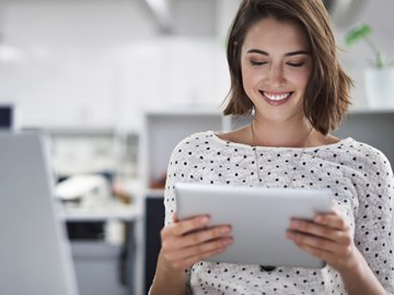 Fragen zu Produktem Eine Frau sitzt im Büro und hält ein Tablet in der Hand, während sie lacht