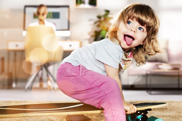 Homeoffice Kind spielt mit einem Skateboard, im Hintergrund die Mutter wie sie im Homeoffice arbeitet