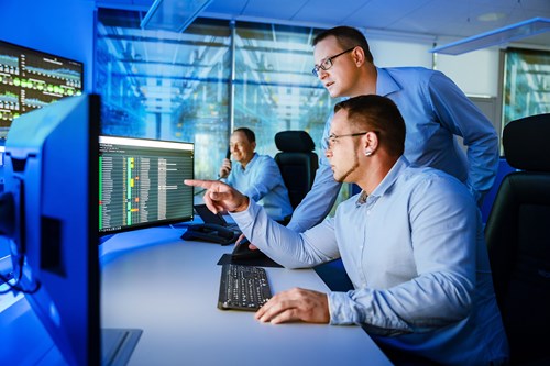 Security-Operation-Center Zwei Männer schauen am Arbeitsplatz auf Monitore, ein dritter sitzt im Hintergrund und telefoniert
