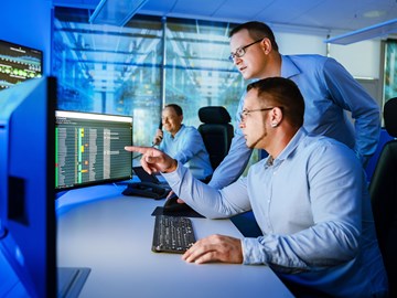 Security-Operation-Center Zwei Männer schauen am Arbeitsplatz auf Monitore, ein dritter sitzt im Hintergrund und telefoniert