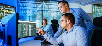 Security-Operation-Center Zwei Männer schauen am Arbeitsplatz auf Monitore, ein dritter sitzt im Hintergrund und telefoniert