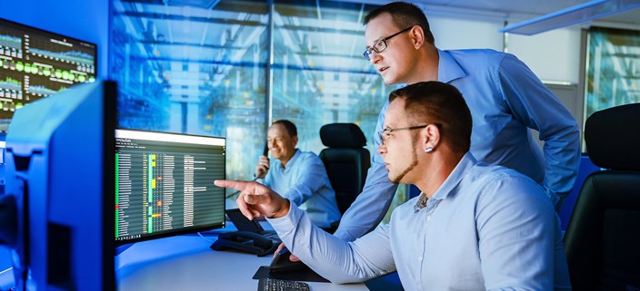 Security-Operation-Center Zwei Männer schauen am Arbeitsplatz auf Monitore, ein dritter sitzt im Hintergrund und telefoniert